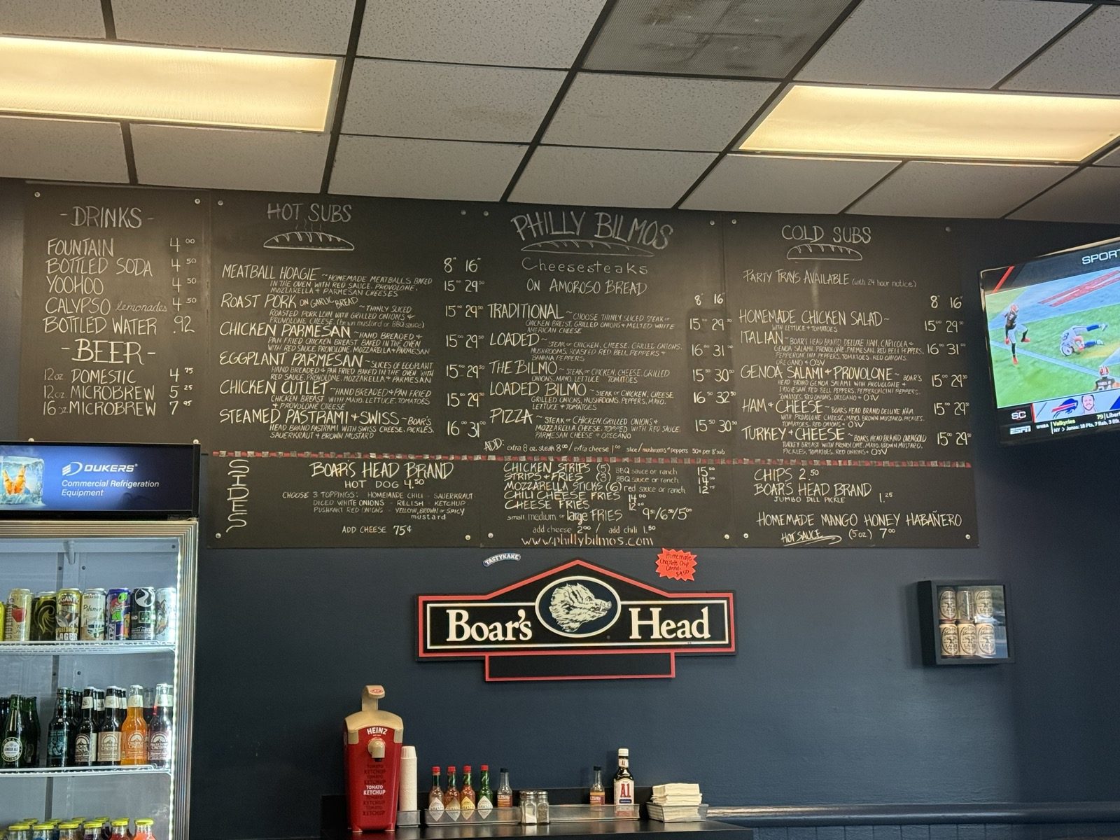 The whole menu, hand-lettered. Cheesesteaks down the middle, cold subs on the right, and a homemade mango honey habanero hot sauce for sale at the bottom of the board.