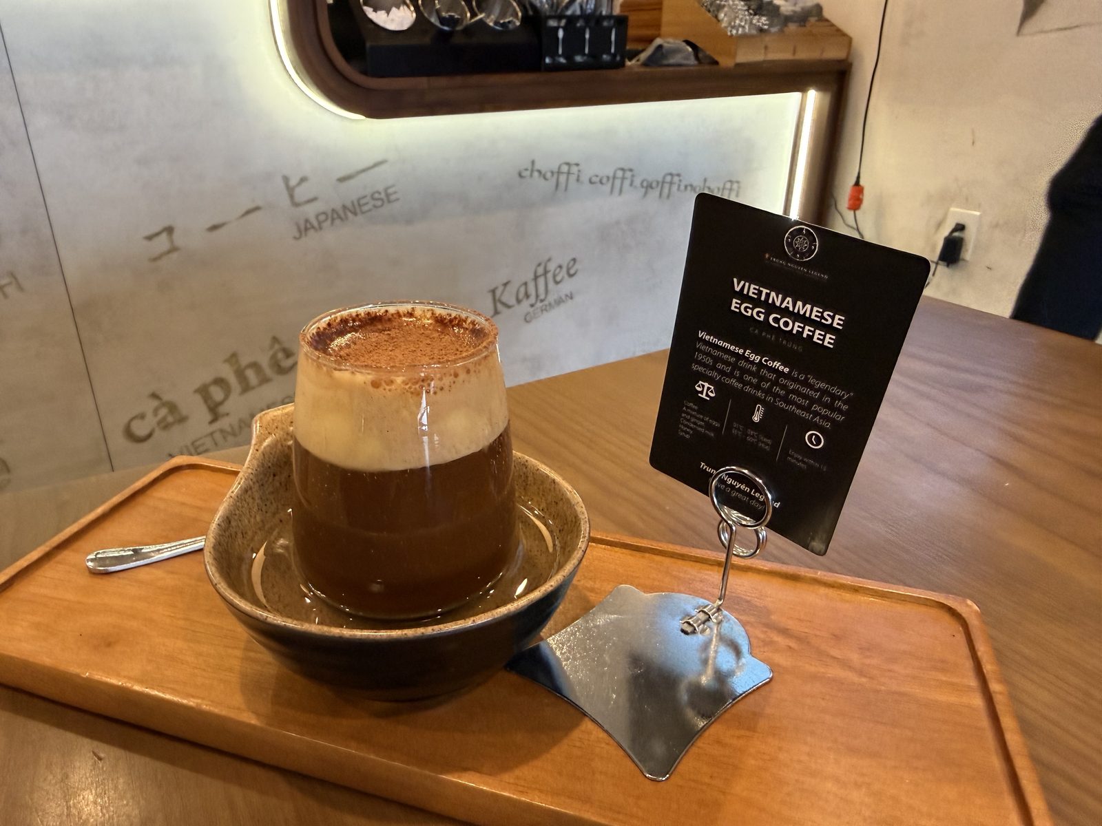 The whole ritual: glass, hot-water bowl, and the card that tells you what you're about to drink, in case the foam didn't already give it away.